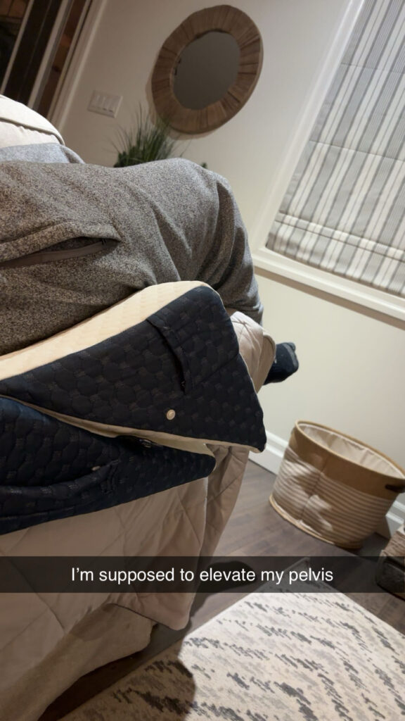 A picture of my legs in grey sweatpants sitting on a wedge shaped pillow with a snapchat caption saying "I'm supposed to elevate my pelvis"